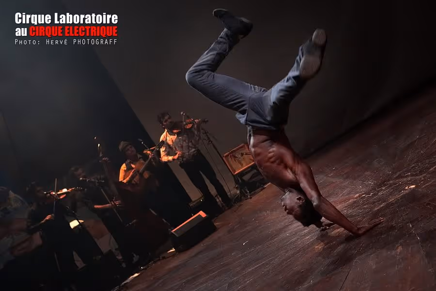 Cirque Électrique Laboratoire Paris acrobate - équilibriste sur les mains, pantalon gris, torse nu, performance scénique nocturne, musiciens live en arrière-plan, spectacle contemporain pluridisciplinaire, photo Hervé Photograff, arts vivants expérimentaux