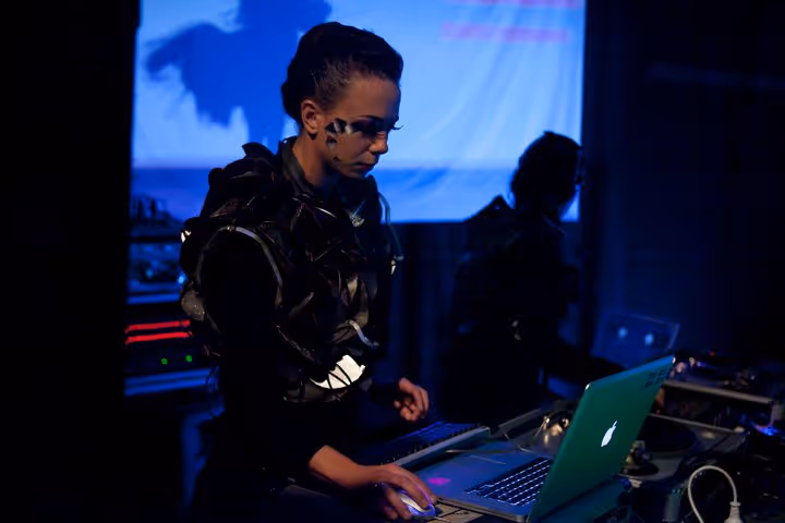 Cirque Électrique laboratoire sonore Paris - DJ femme avec maquillage artistique et veste militaire, équipement électronique, ordinateur Apple, table de mixage, création musicale live, performance audiovisuelle nocturne, esthétique futuriste