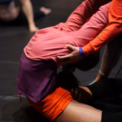 Deux femmes en tenue colorée, l'une aidant l'autre à s'étirer assise au sol dans une salle d'entraînement.