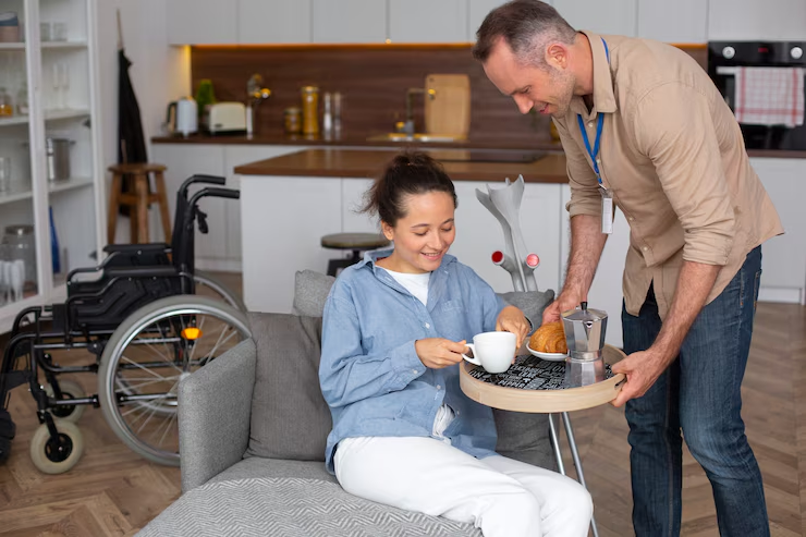 Caregiver serving tea to person in blue shirt near wheelchair at home
