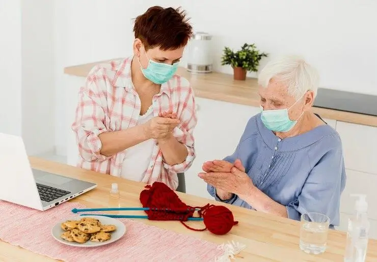 Two people with masks knitting together, with cookies and laptop nearby