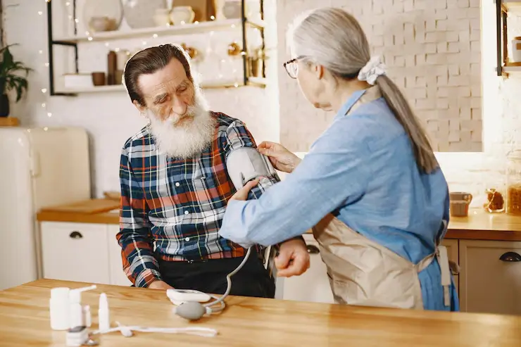Elderly couple in kitchen, one helping the other with medical device