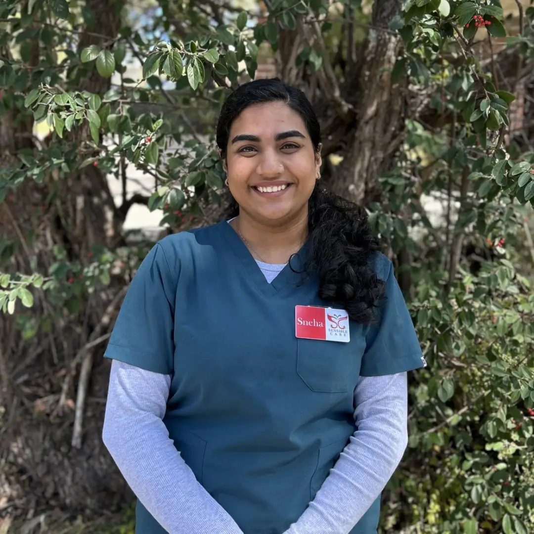 Smiling healthcare worker in blue scrubs standing in front of green foliage
