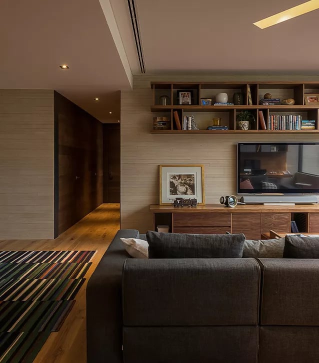 Modern living room with gray sofa facing a wooden TV console, wall-mounted shelves with books and decor, and wooden flooring with a striped rug.