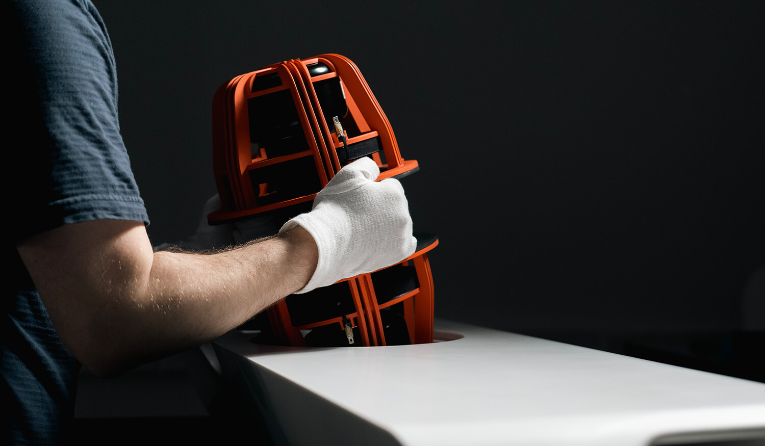 Person wearing a white glove handling an orange and black surveying instrument next to a white surface against a dark background.