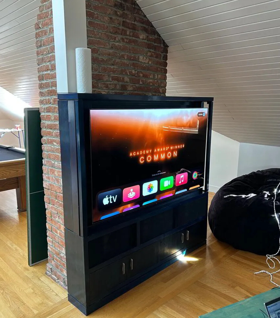 Flat-screen TV mounted on a black stand against a brick column in a living room, displaying an Apple TV interface with the text 'Academy Award Winner COMMON'.