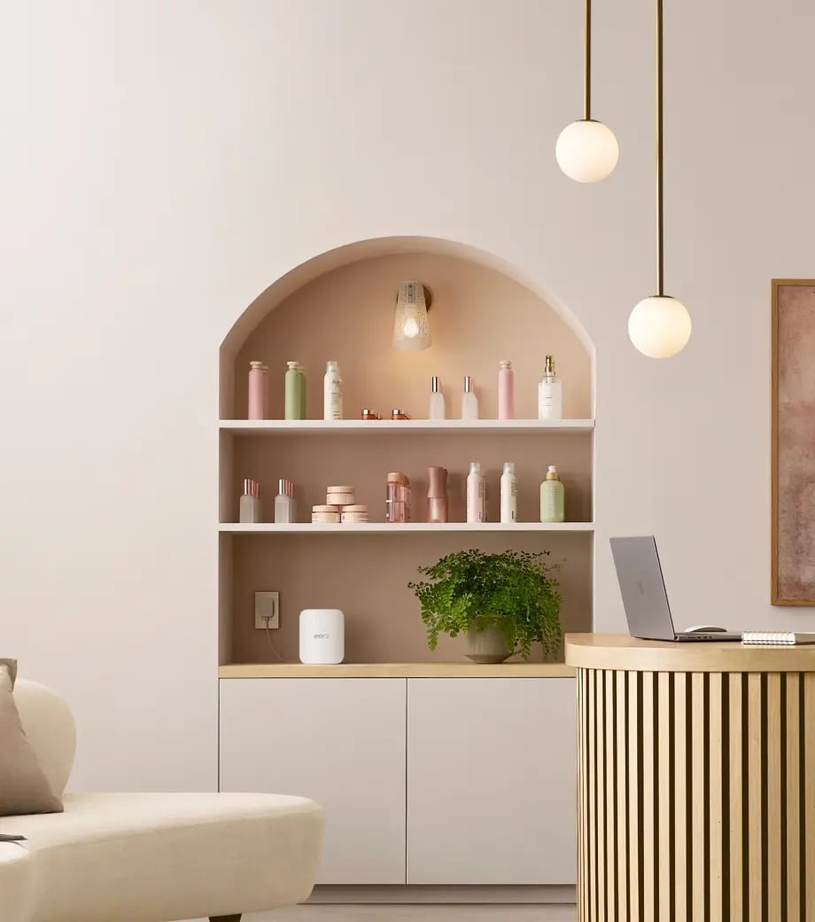 Minimalist interior with wooden slatted round desk holding a laptop and a white cabinet with shelves displaying skincare bottles and a green potted plant.