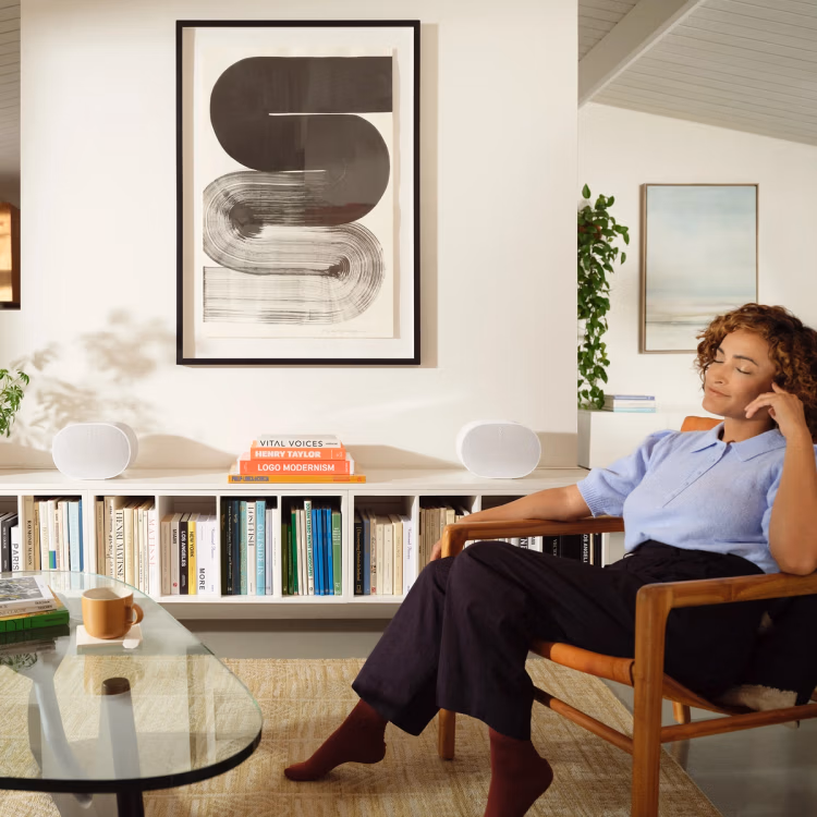 Woman in a light blue shirt and black pants sitting in a wooden chair in a modern living room with a bookshelf, abstract wall art, and glass coffee table with a mug.