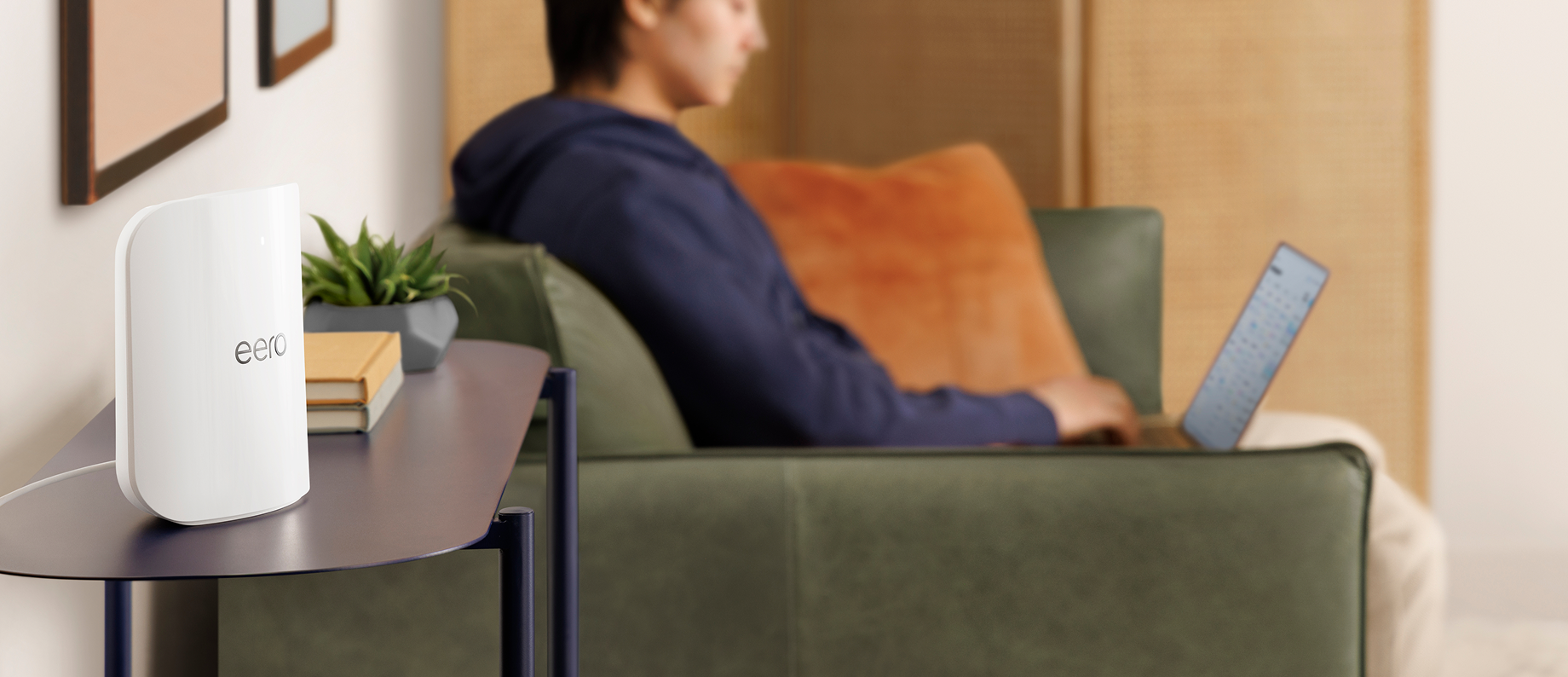 White eero Wi-Fi router on a side table next to a plant and books, with a person using a laptop on a green sofa in the background.