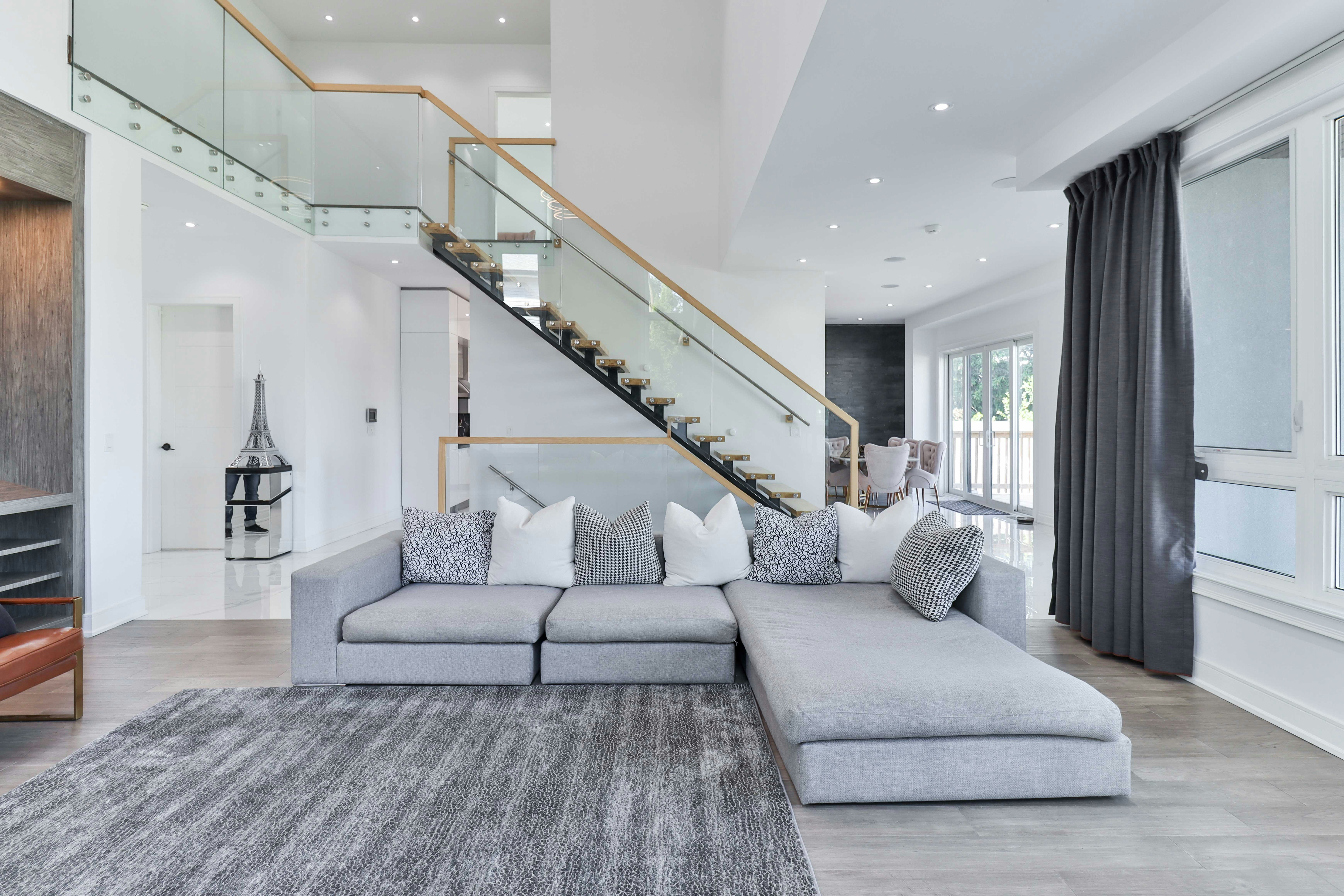 Modern living room with a gray sectional sofa, patterned pillows, glass staircase with wood railing, and large windows with dark curtains.