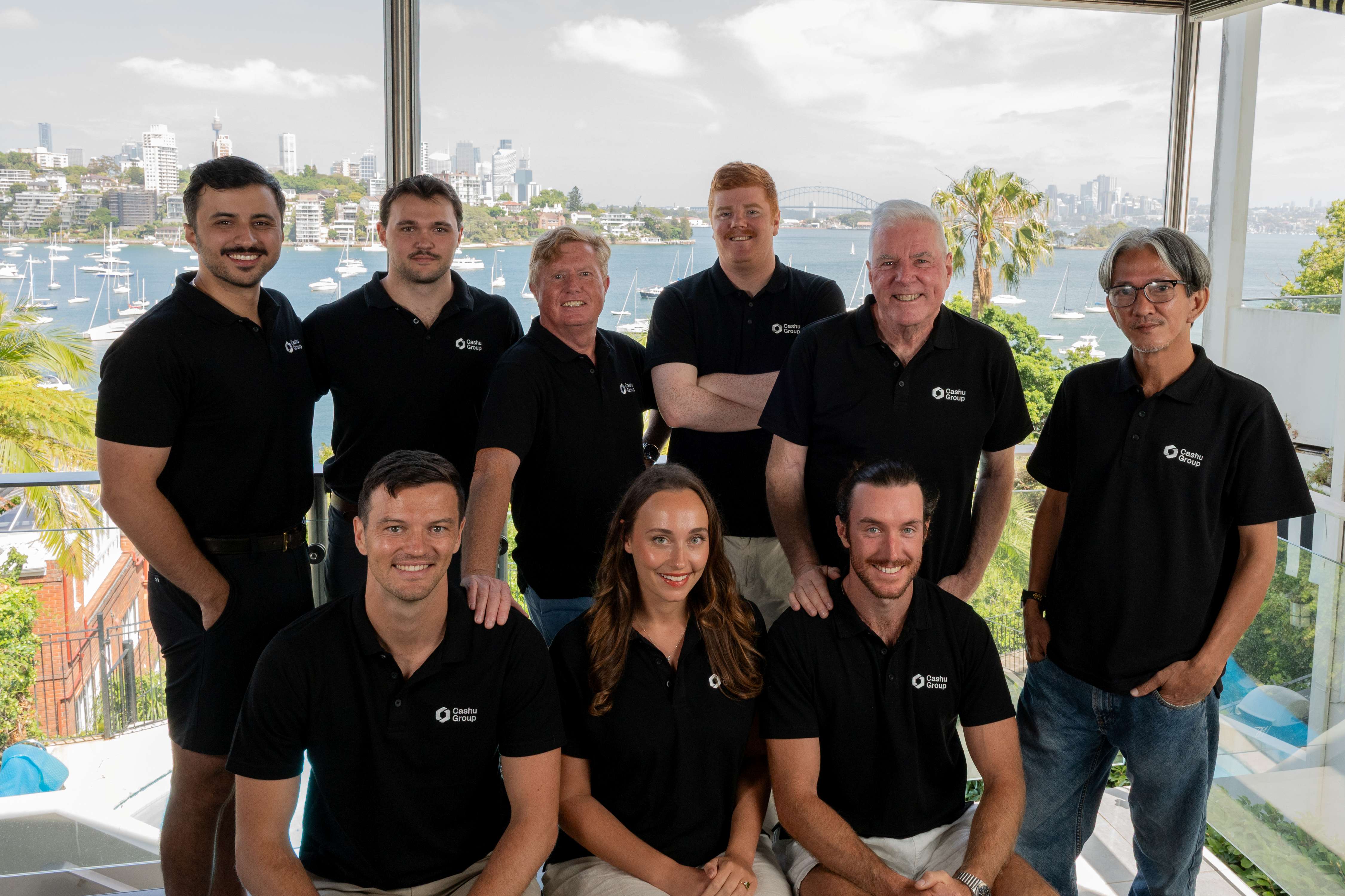 Cash Group Team  of nine people wearing black Cashu Group polo shirts posing indoors with a bay and city skyline in the background.