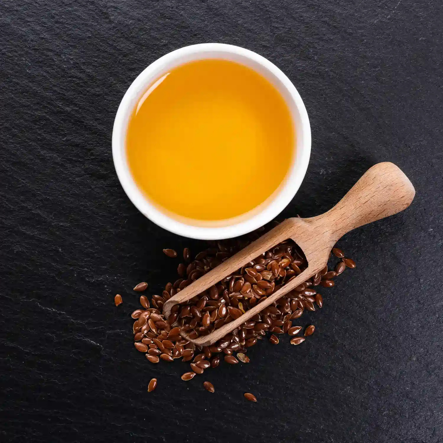 Golden Flaxseed Oil in a small white bowl, with a wooden scoop with Flaxseeds