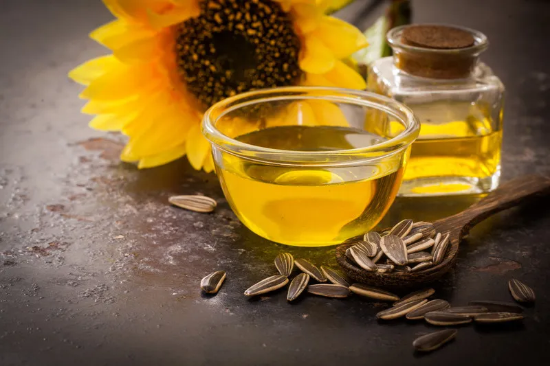 Sunflower oil in small glass bowl with sunflower seeds in the foreground and sunflower flower in the background