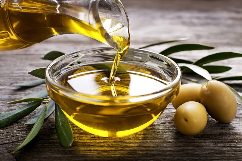 Olive oil being poured into a small glass bowl with green olives on the side