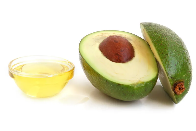 Small glass bowl of avocado oil with a halved avocado beside it on a white background