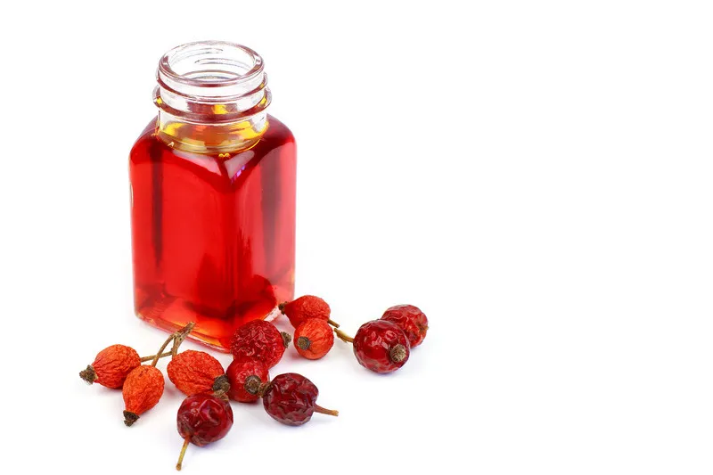 Rosehip oil in small glass jar with dried rosehip buds in the foreground on a white background