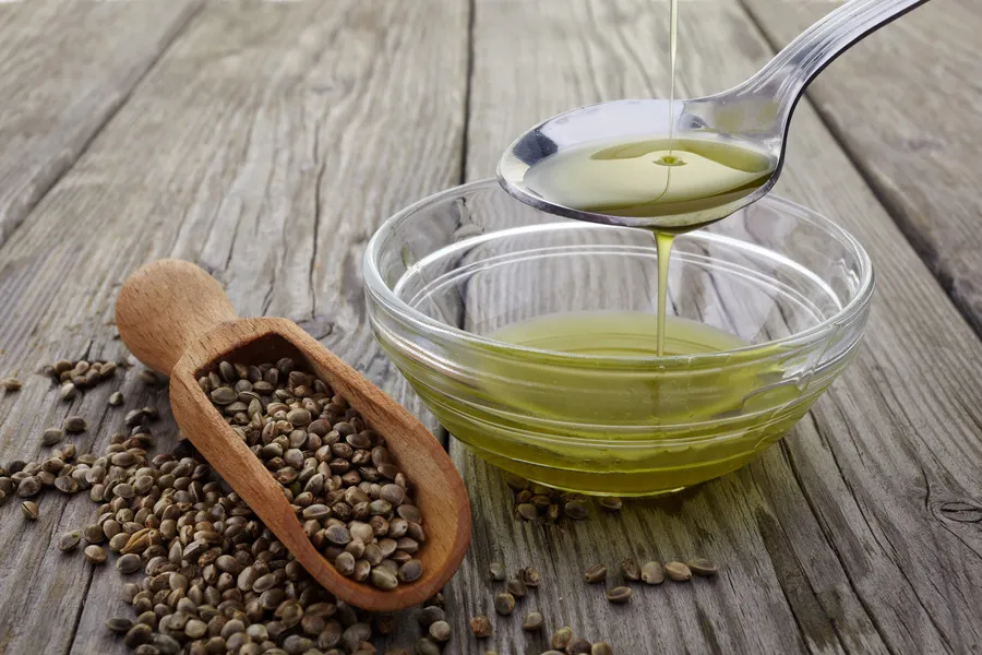 Hemp seed oil being pouring onto a spoon, flowing into a small glass bowl with a wooden scoop with hemp seeds beside it
