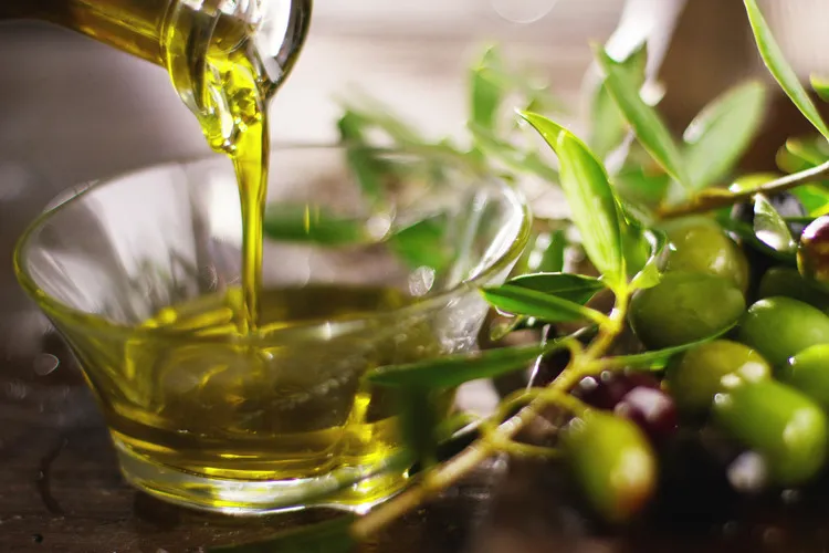 Olive oil being poured into small glass bowl with green olives beside it