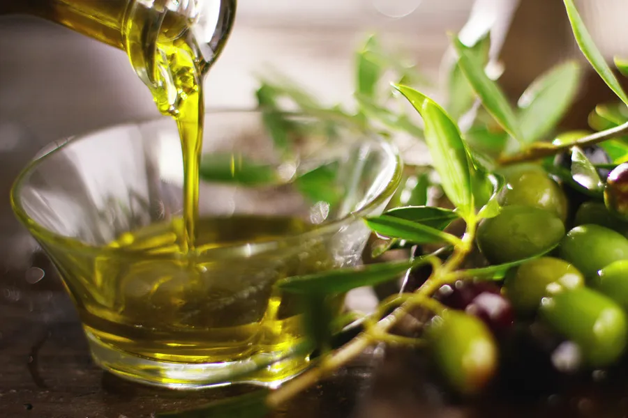 Olive oil being poured into small glass bowl with green olives beside it