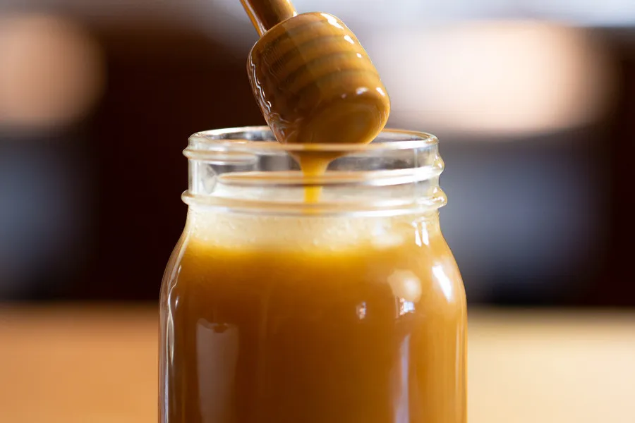 Honey dipper drizzling premium Mānuka honey into a glass jar.