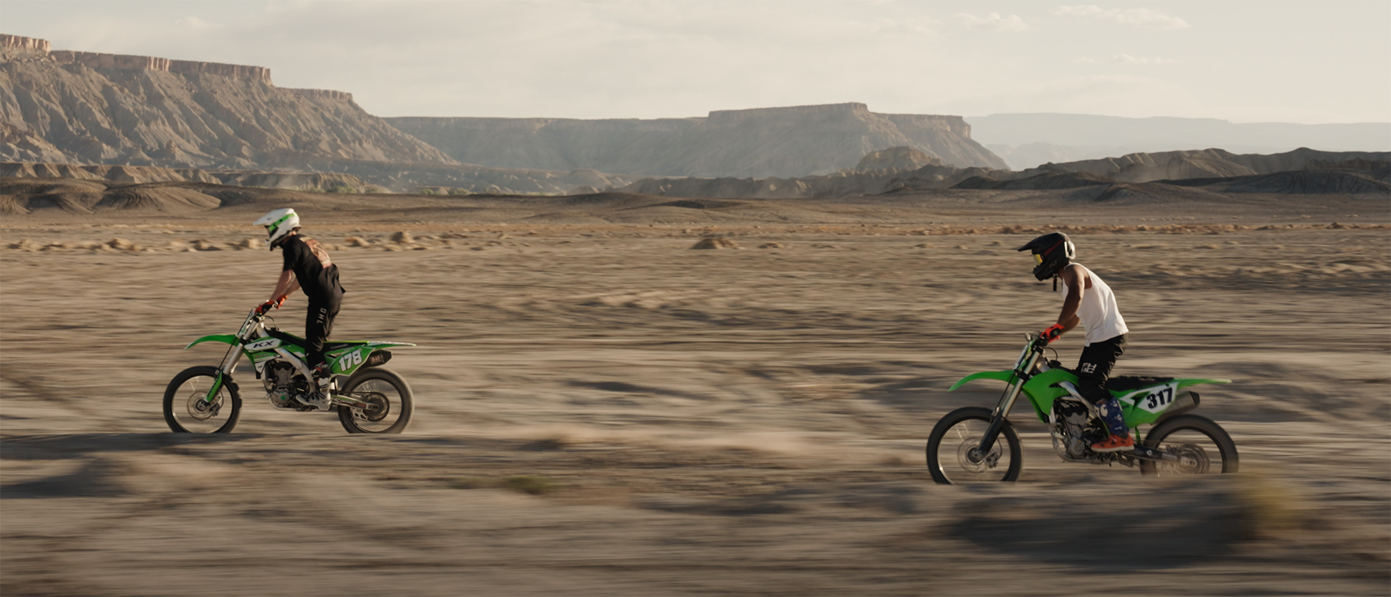 Two motocross riders in helmets riding green dirt bikes across a flat desert landscape with rocky mesas in the background - from Leo Sideras' project 'Swingarm City Takeover.'