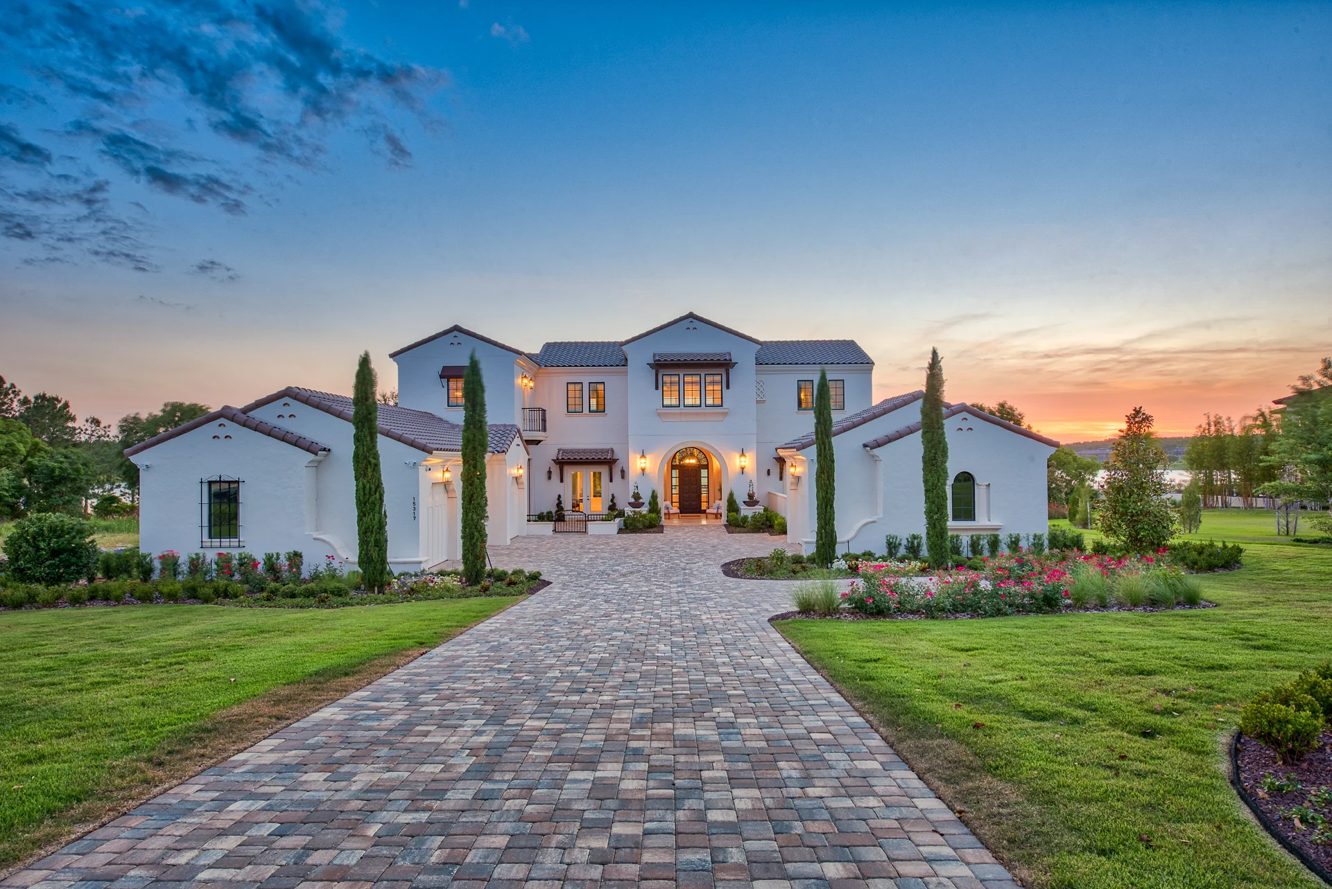 Large white Mediterranean-style house with tiled roof and tall cypress trees along a stone driveway at sunset.