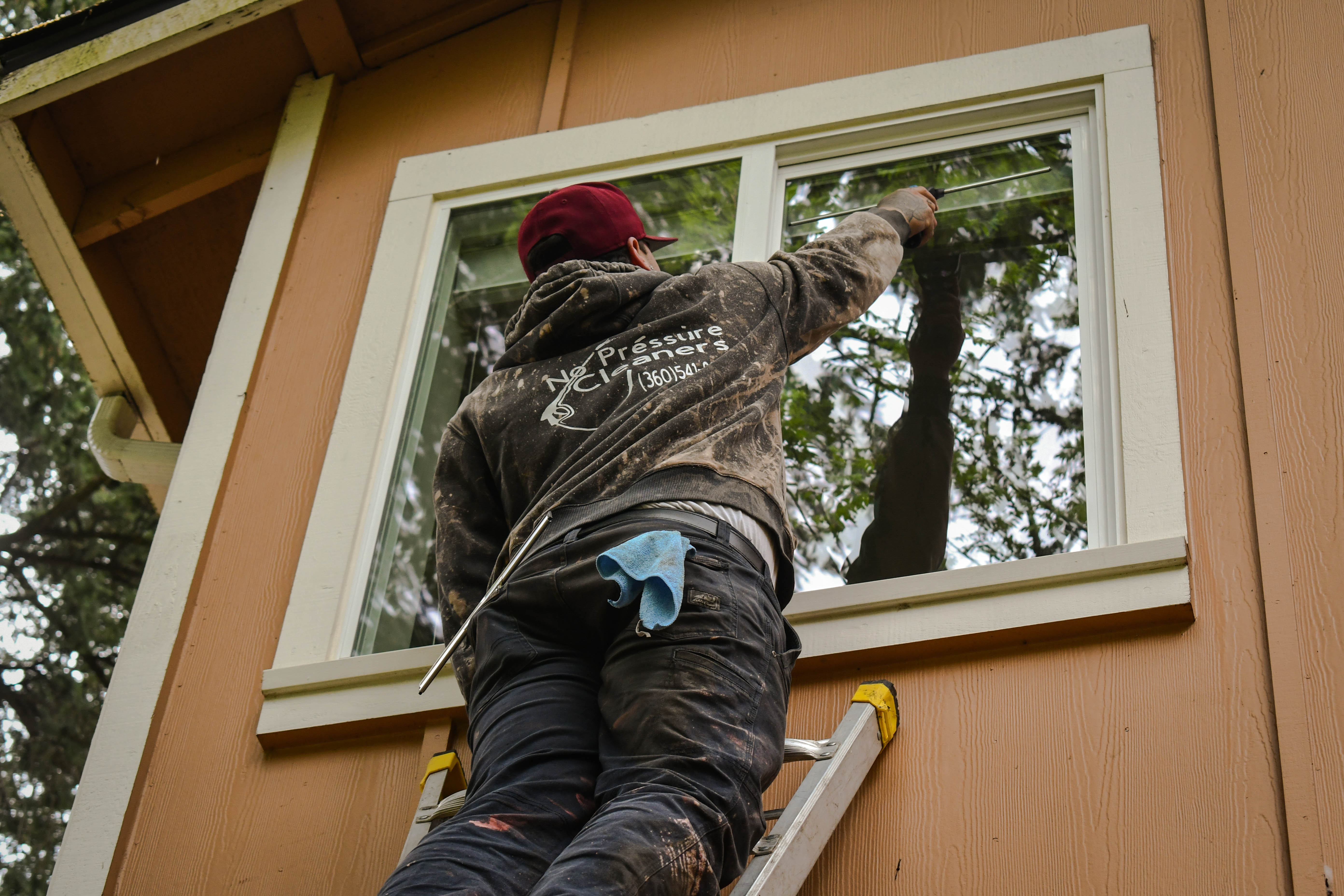 A dirty worker in a maroon cap and hoodie cleaning an outdoor window while standing on a ladder.