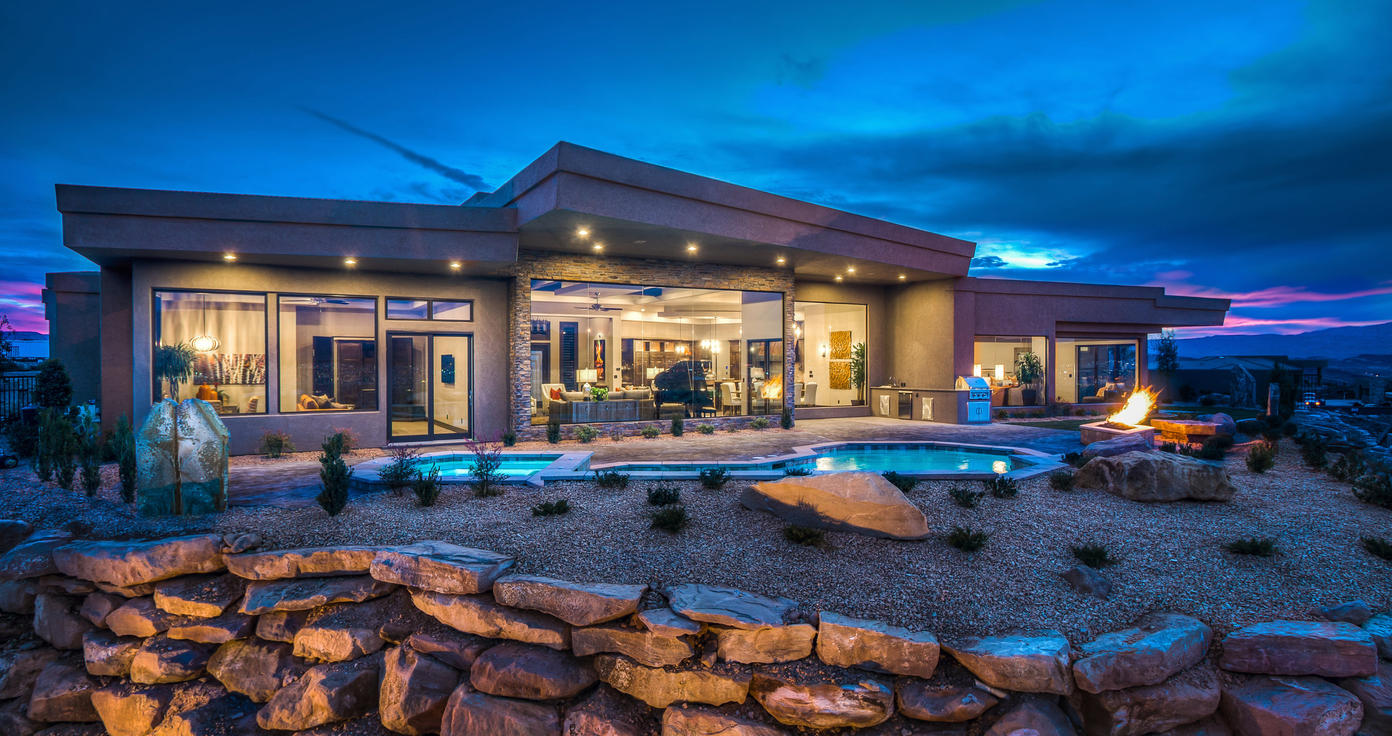Modern single-story house at dusk with large glass windows, outdoor pool, fire pit, and stone landscaping.