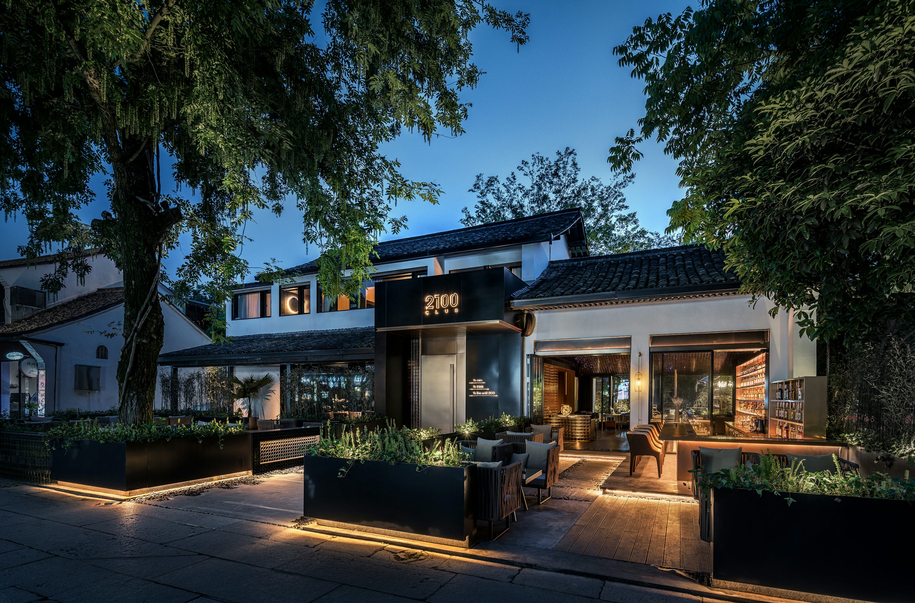 Outdoor seating area of a modern restaurant with illuminated planters and trees at dusk.