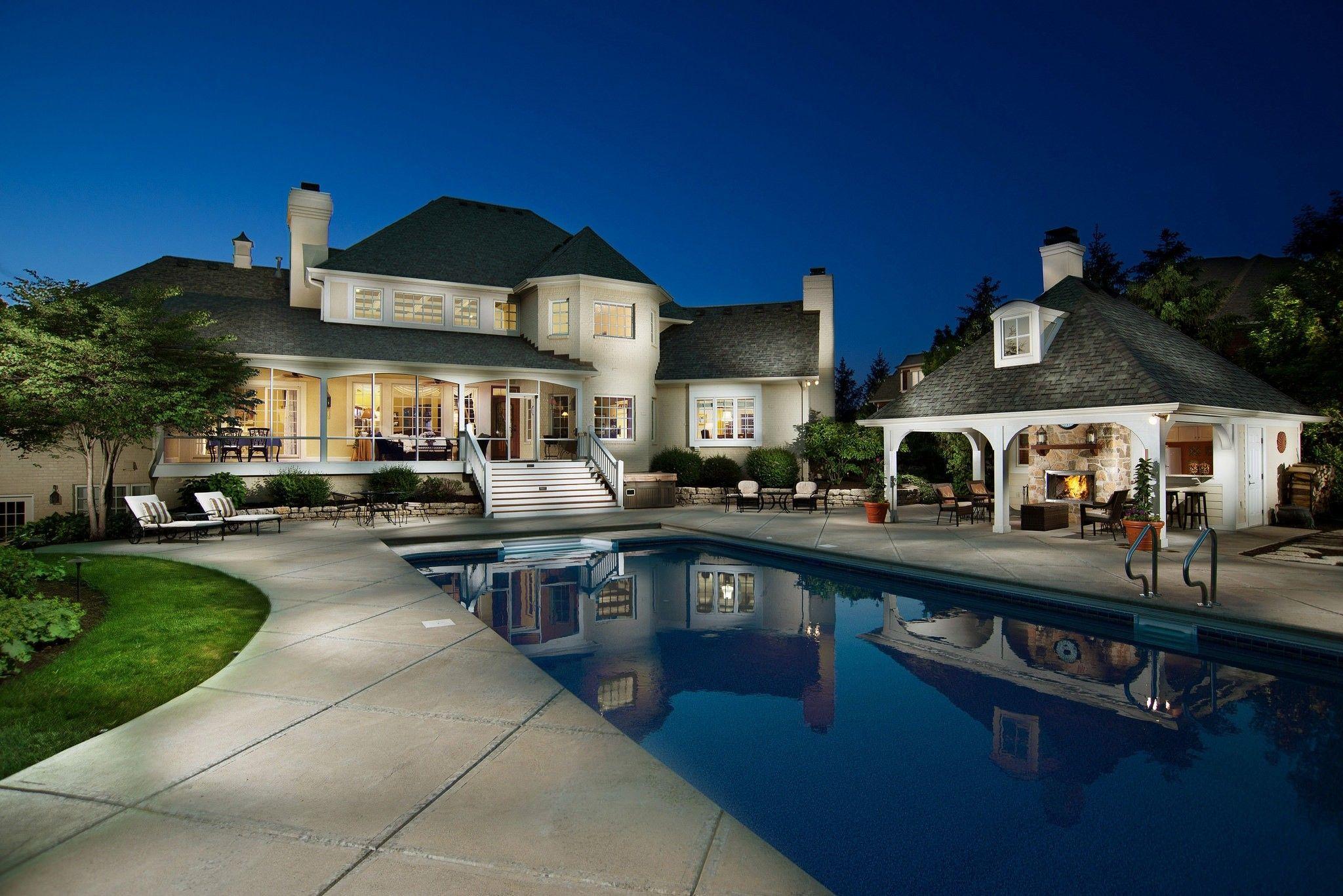 Large illuminated house with a covered patio, outdoor fireplace, and a swimming pool reflecting the building at dusk.