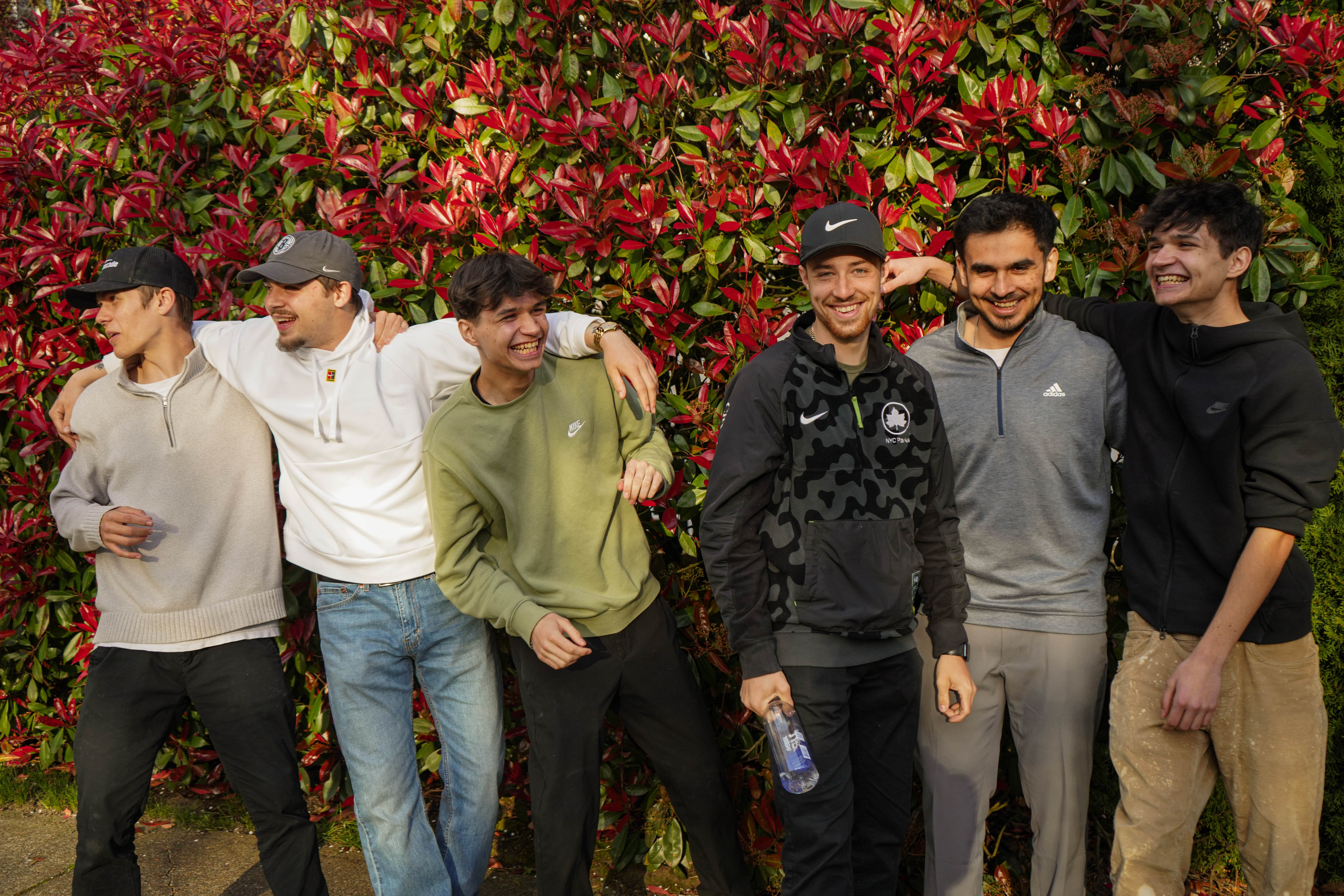 Six young men standing closely in front of vibrant red and green foliage, smiling and laughing together.