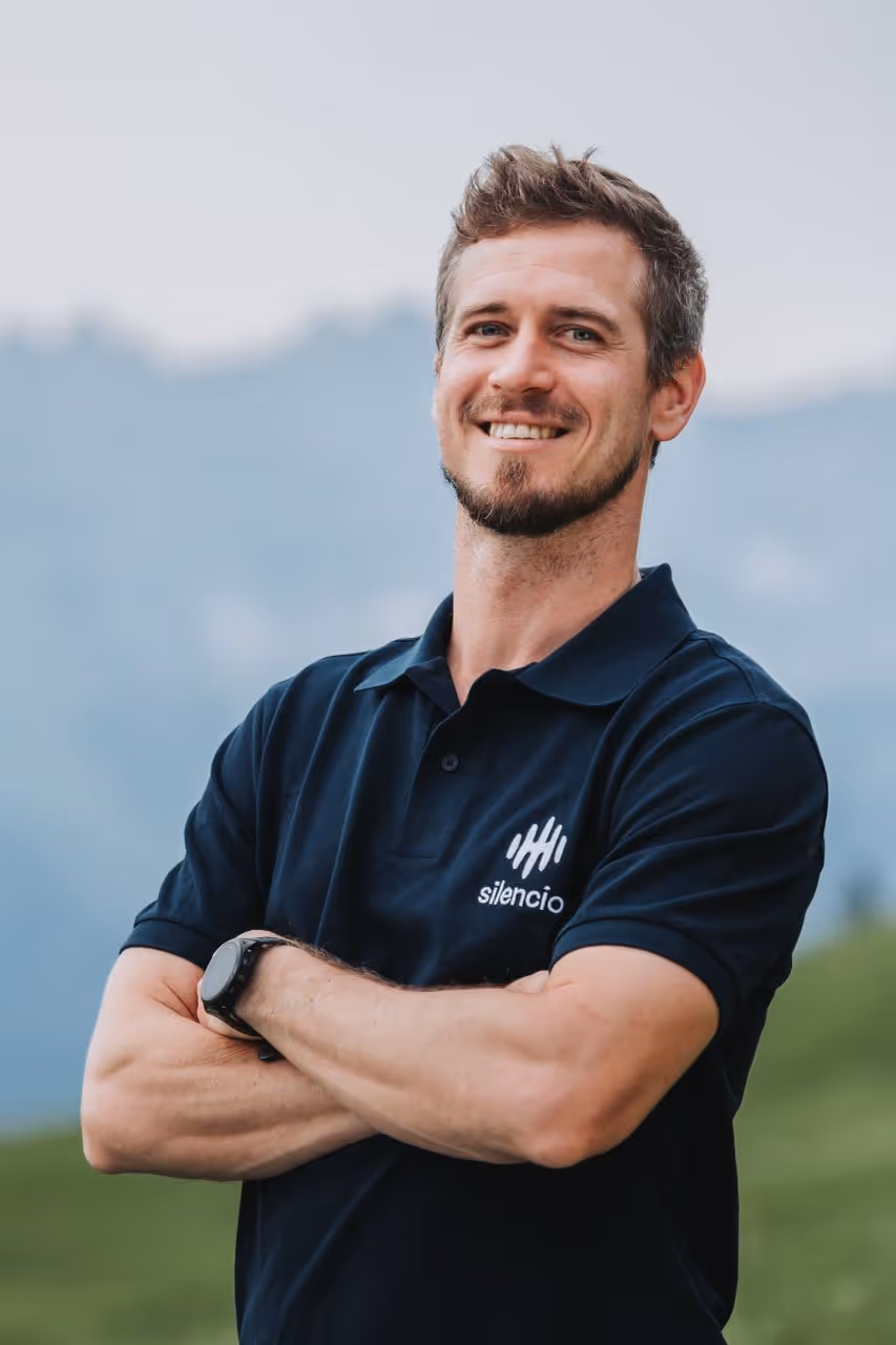Man in polo shirt with arms crossed, smiling outdoors.