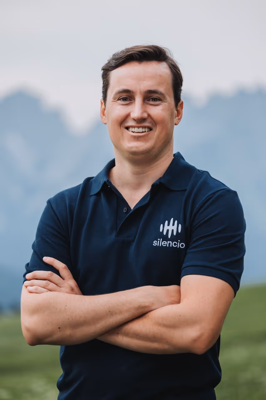 Man in polo shirt with arms crossed, mountains in background.