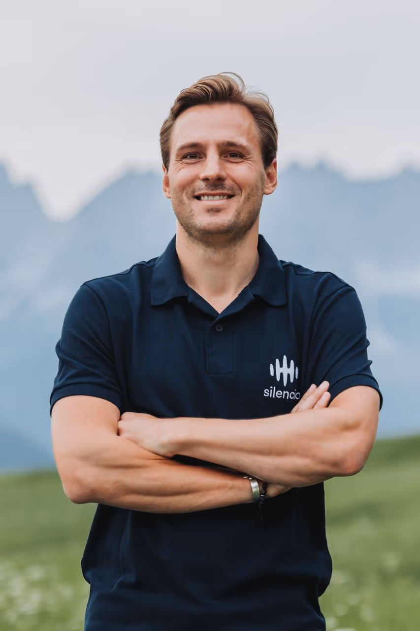Man in polo shirt with arms crossed, mountains in background.
