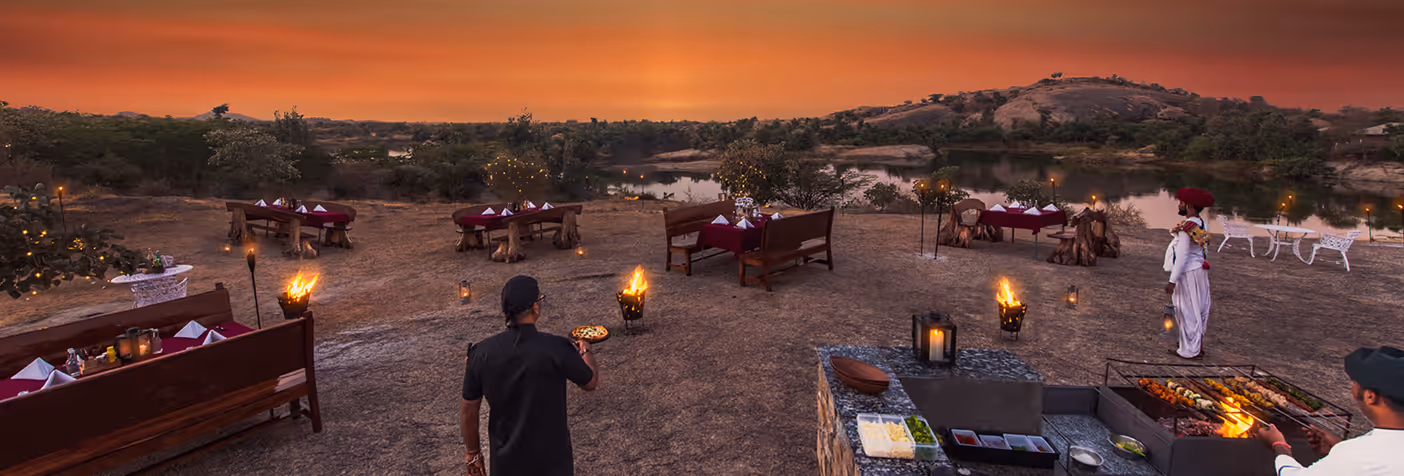 Outdoor dining setup at sunset with tables, benches, torches, and a chef grilling skewers beside a lake and rocky hills.