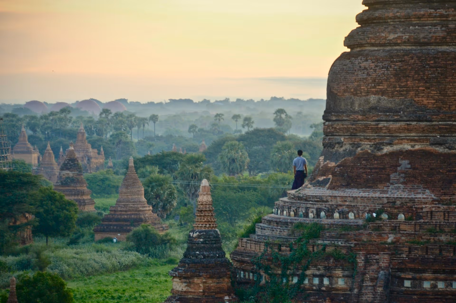 Person standing on an ancient brick temple overlooking a lush green landscape dotted with multiple pagodas at sunrise.