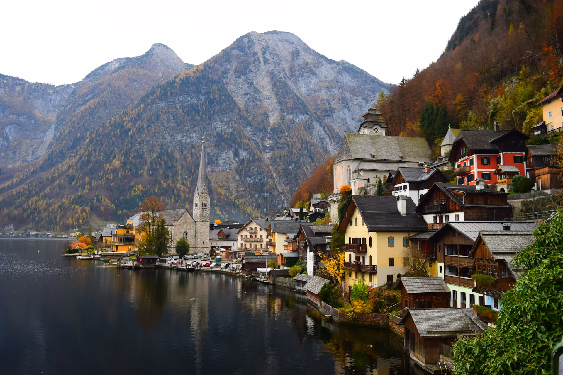 Picturesque village with colorful houses and a church spire beside a calm lake, backed by tall forested mountains in autumn.