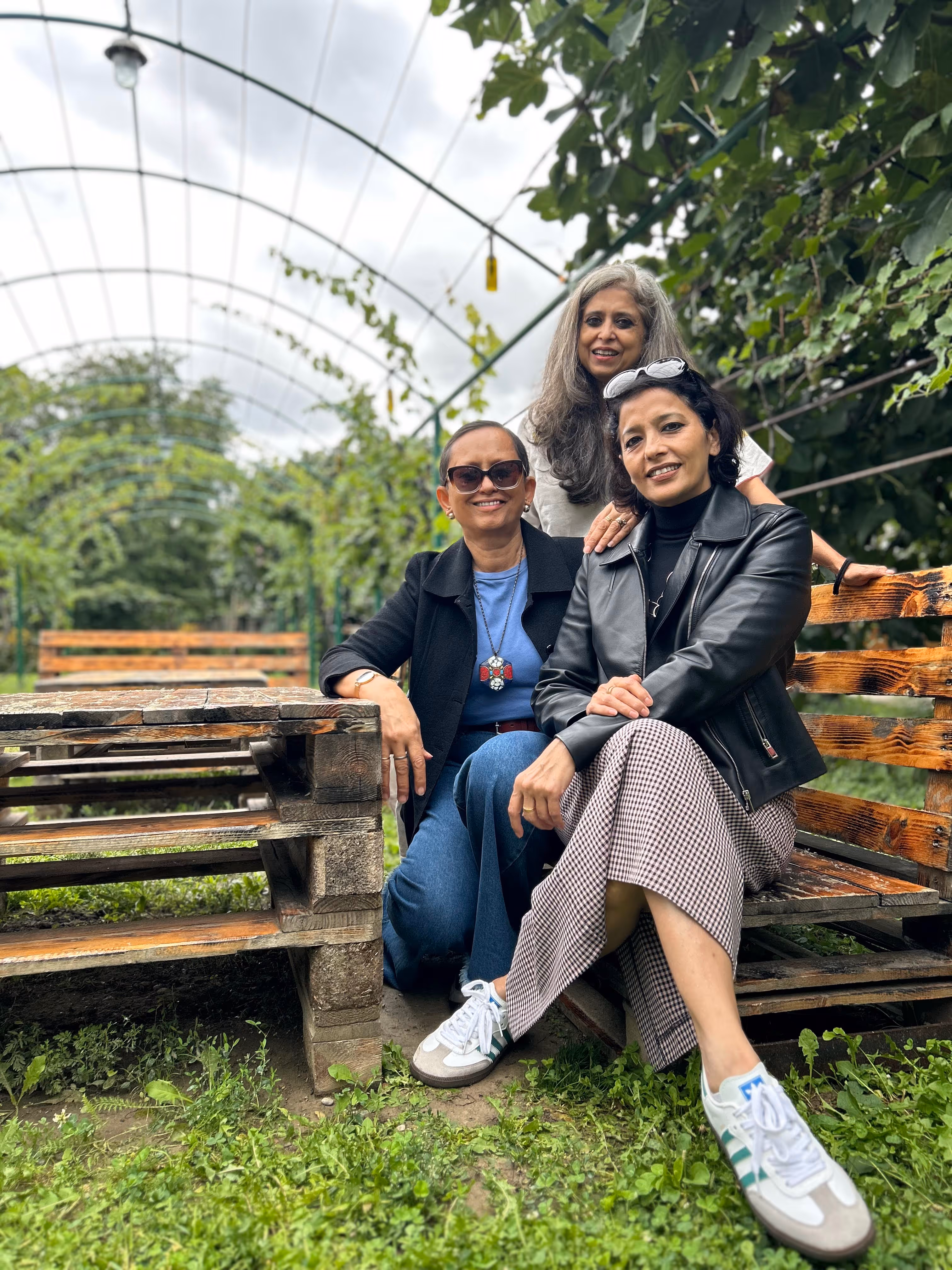 Three women smiling and sitting on and beside a rustic wooden bench in a vine-covered outdoor garden structure.