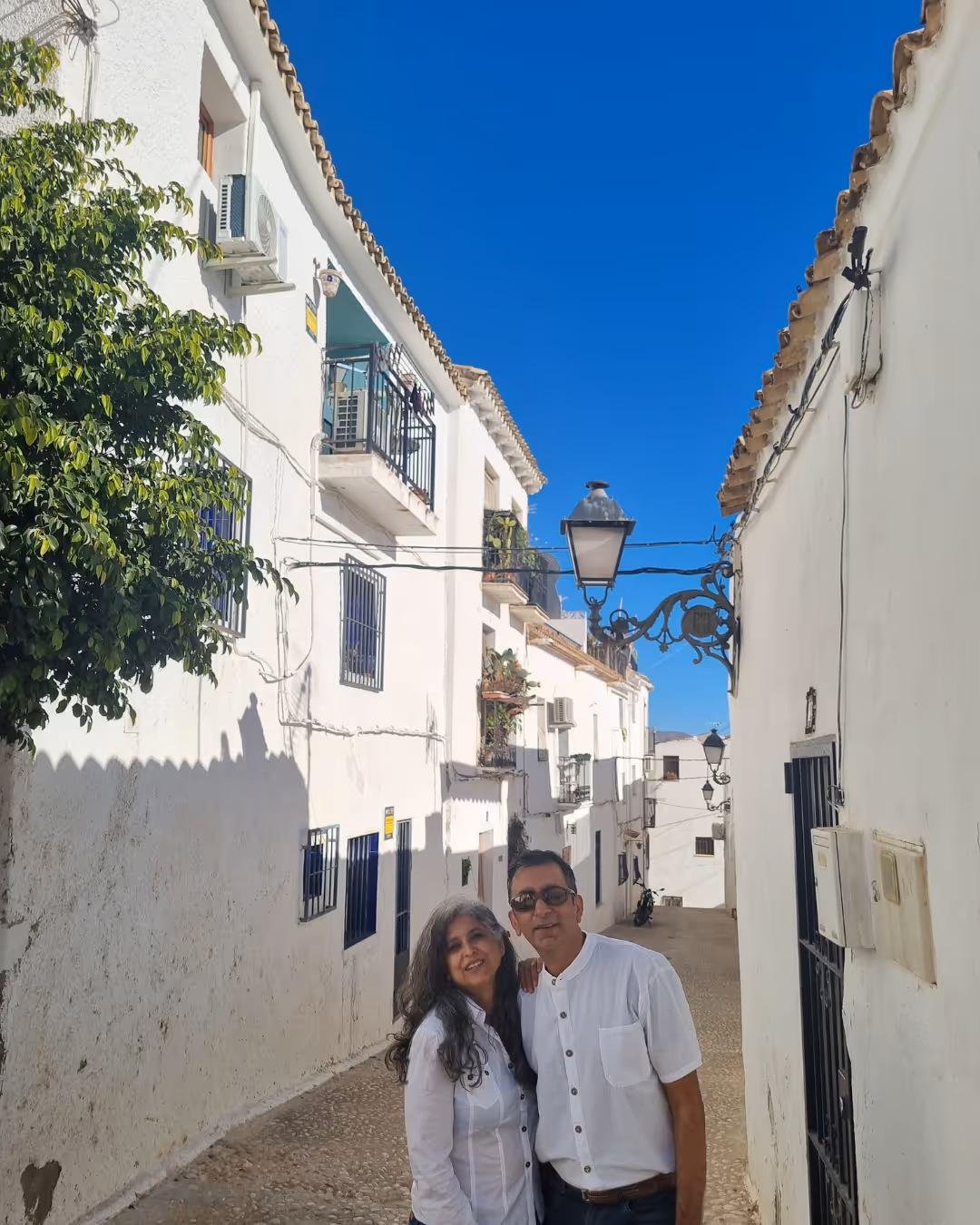 Couple smiling and standing closely on a narrow cobblestone street lined with white buildings under a bright blue sky.