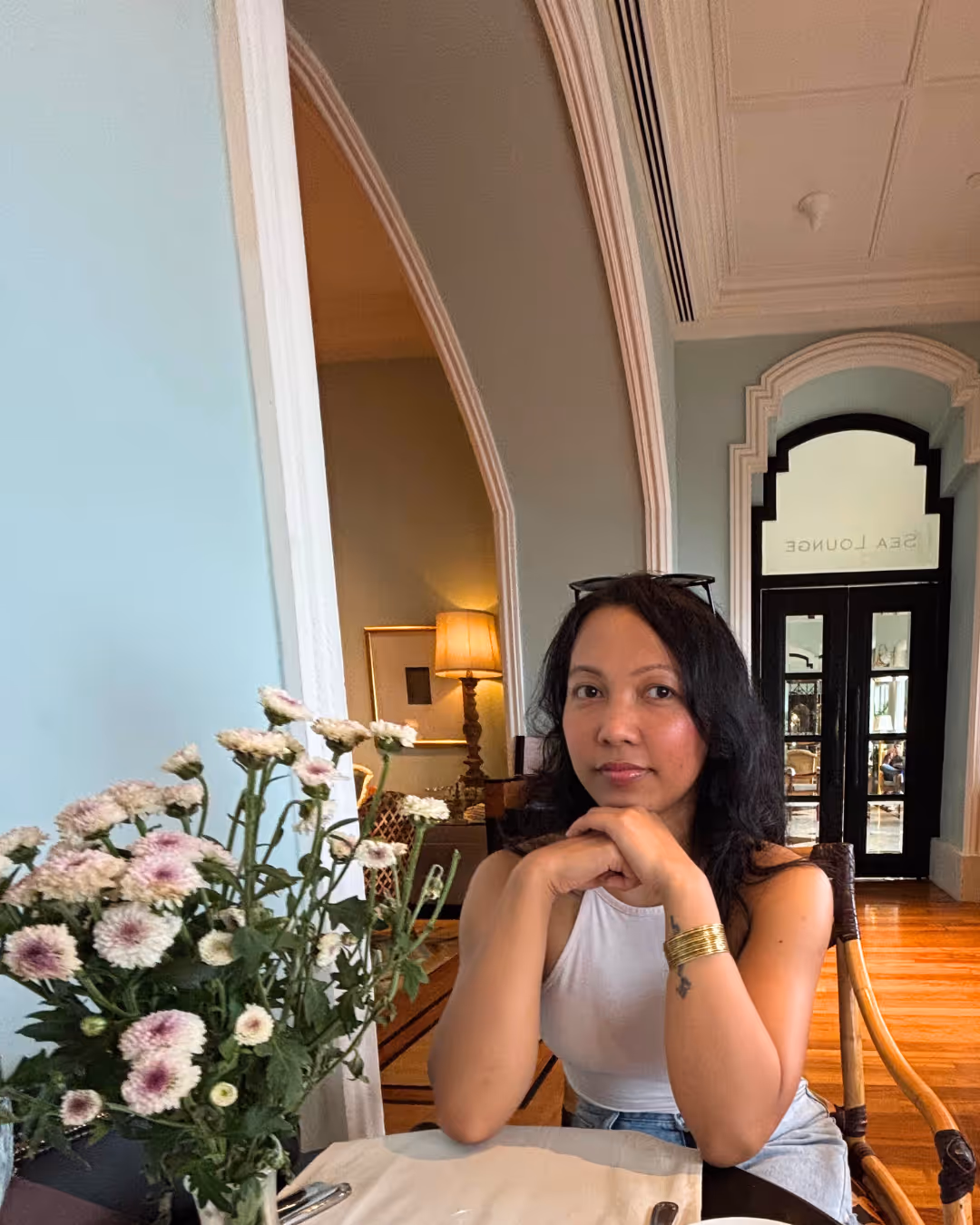 Woman sitting at a table with folded hands in a cozy room with arched doorways and a vase of flowers in the foreground.