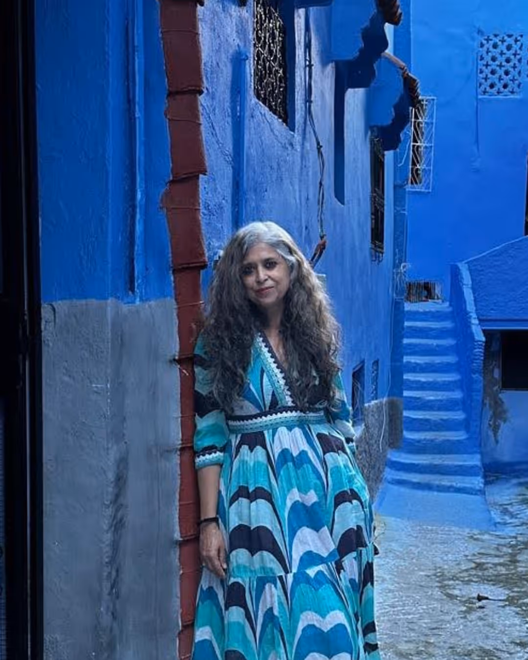 Woman with long gray hair wearing a blue patterned dress leans against a blue and gray wall in a narrow blue-painted alley with stairs.