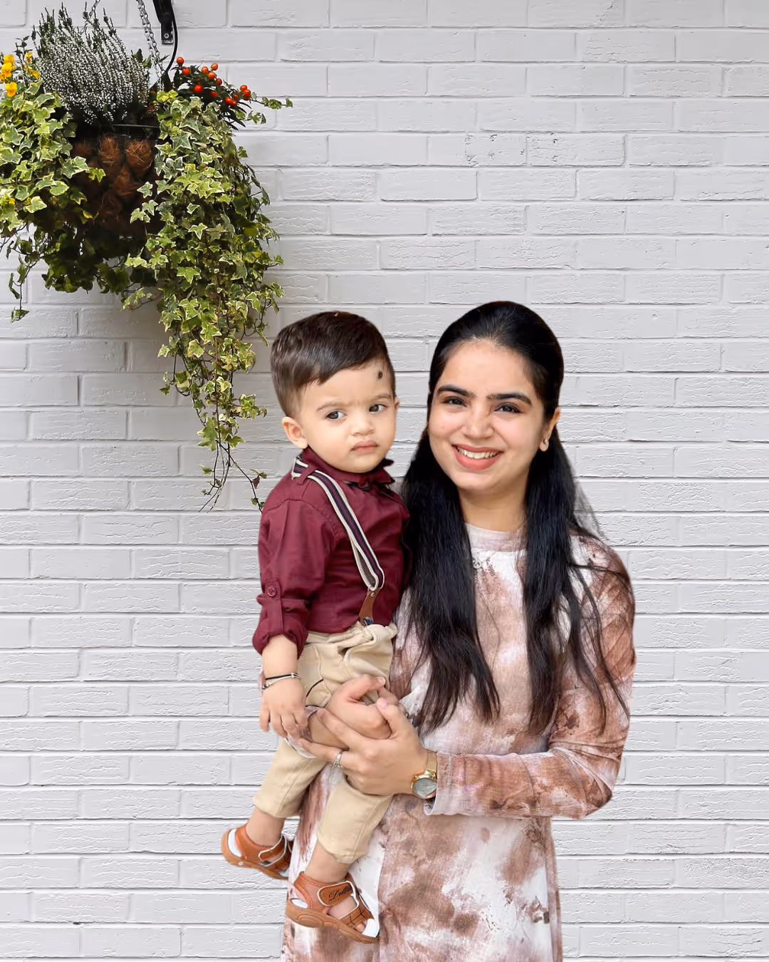 Woman with long dark hair holding a young boy dressed in a maroon shirt and beige pants, standing in front of a white brick wall with a hanging green plant.