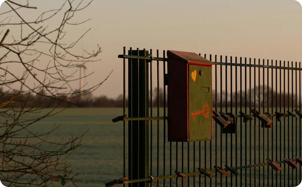 Green metal fence with multiple locks attached and a green box featuring a red key and a yellow heart, set against a field at sunset.