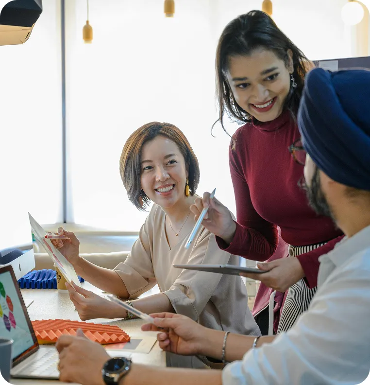 Three diverse coworkers smiling and collaborating with documents and a laptop in a bright office.