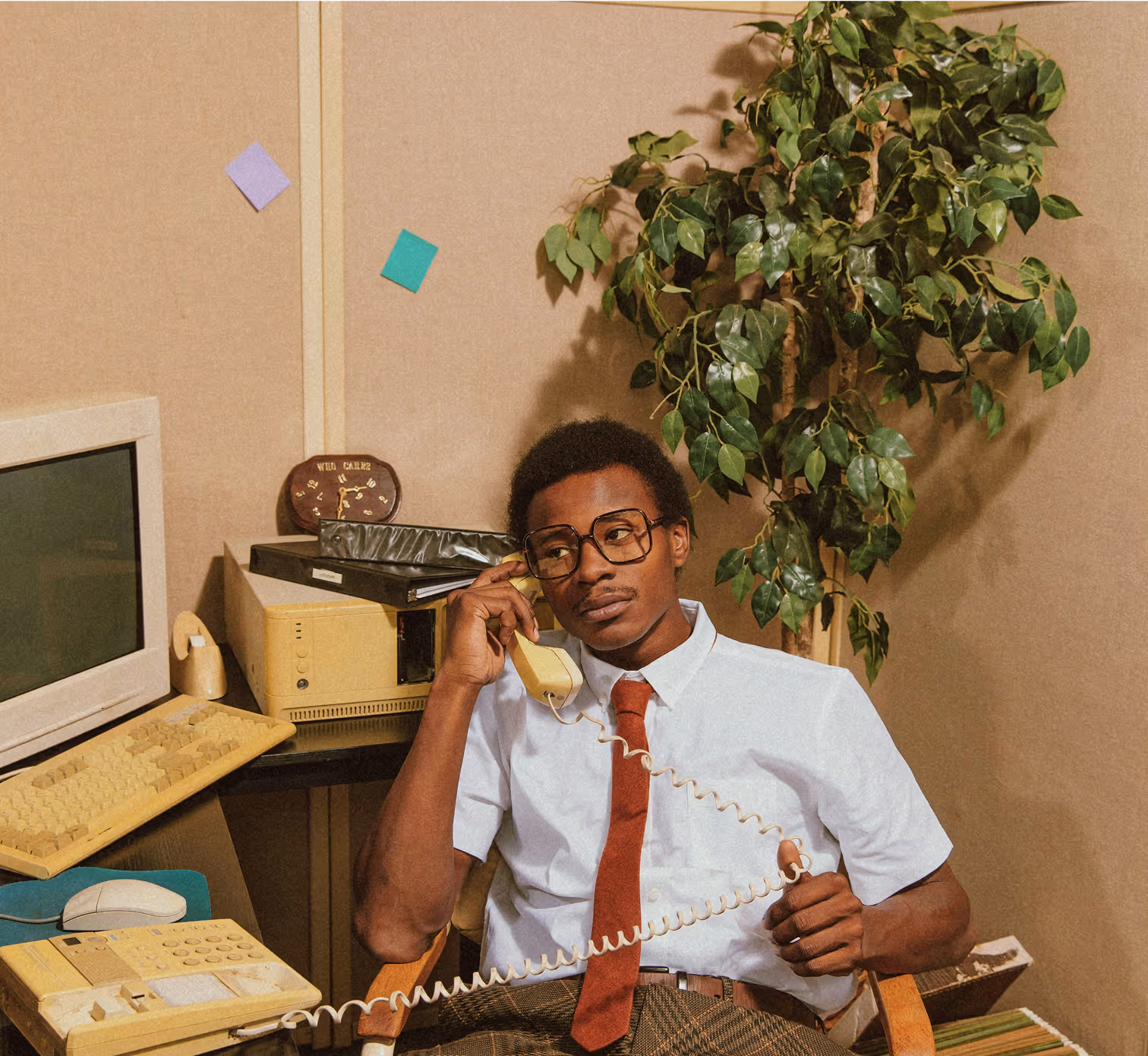 Un homme en chemise et cravate assis à un bureau rétro des années 80, parlant au téléphone filaire avec un ordinateur ancien à côté de lui.