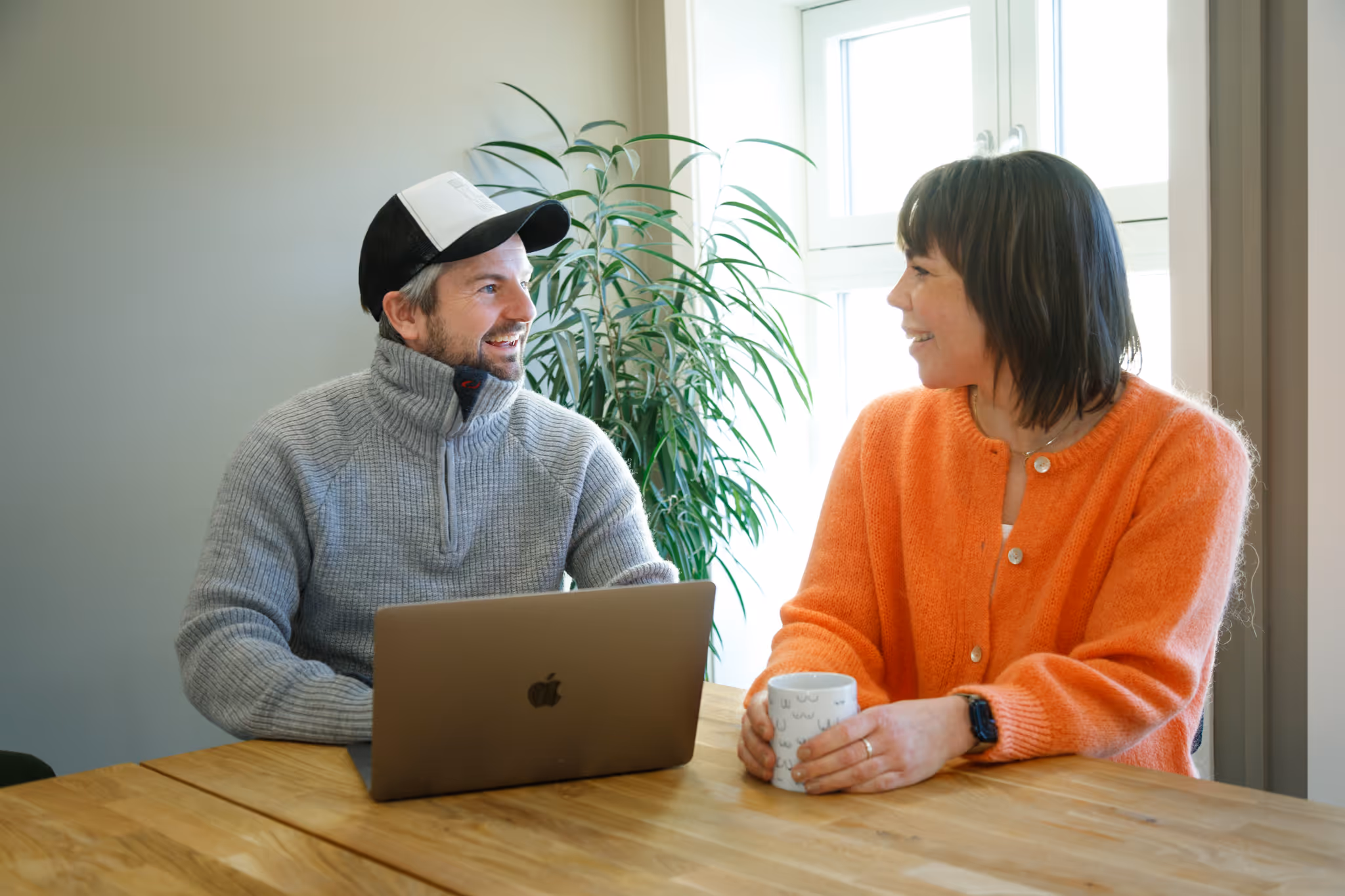 To personer som smiler og snakker ved et trebord, en mann med grå genser og caps ved en bærbar datamaskin, og en kvinne i oransje genser som holder en kopp.