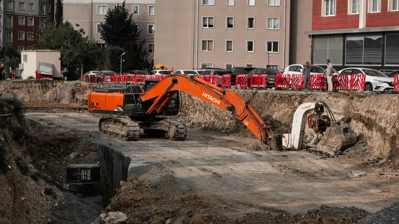 Oranžový bagr Hitachi kope na staveništi s hlubokým výkopem a dva dělníci hovoří u červených zábran.