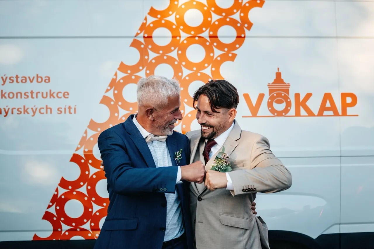 Two men in suits smiling and bumping fists in front of a vehicle with an orange circular pattern and the word VOKAP.