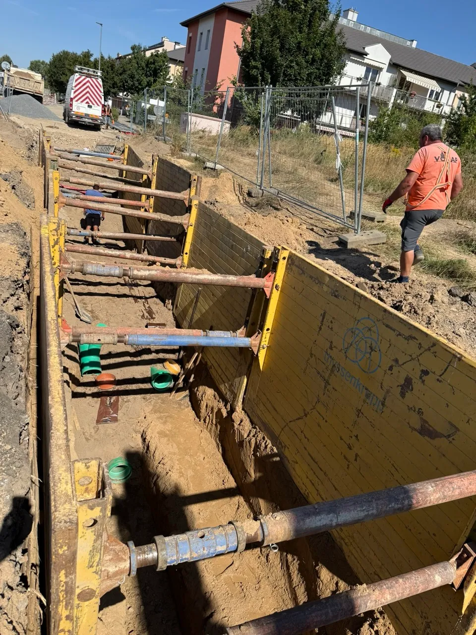 Staveniště s velkým výkopem pro inženýrské sítě, pracovník v oranžovém tričku a dodávka na pozadí.