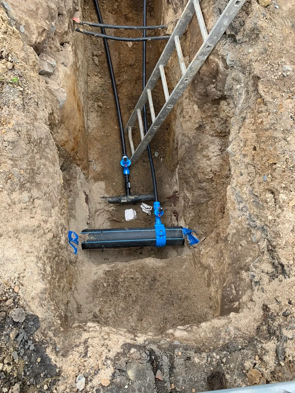 Výkop s uloženými černými plastovými trubkami spojenými modrými spojkami a kovovým žebříkem v zemi.